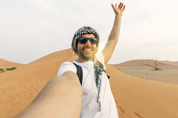 mladý muž v bílém tričku v poušti, treveling v SAE na safari, nosí klobouk a batoh, objevování přírody písečné krásy. dělat selfie v pozadí západu slunce - Fotografie, Obrázek
