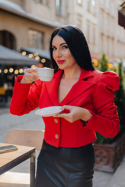 Женщина с красными полными губами и темными волосами держит чашку кофе, стоя на террасе открытого кафе. Стильная леди в черной юбке и красной куртке наслаждается горячим напитком в свободное время. - Фото, изображение