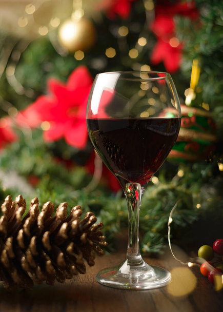 木のテーブルに赤ワインのガラスとクリスマスの装飾. 背景にあるクリスマスツリーとライト. - 写真・画像