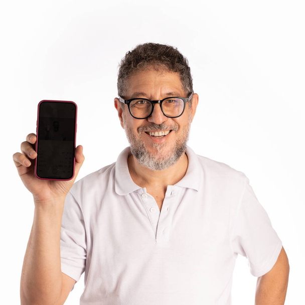 Un hombre alegre de cincuenta años de edad, con pelo rizado, barba y gafas, muestra con confianza una pantalla de teléfono inteligente, aislado en un fondo de estudio blanco. Lleva una camisa de polo y mira directamente a la cámara.. - Foto, Imagen