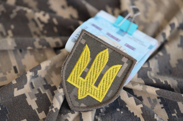 Simbolo dell'esercito ucraino e un mucchio di fatture grivna sull'uniforme militare. Pagamenti ai soldati dell'esercito ucraino, stipendi ai militari. Guerra in Ucraina - Foto, immagini