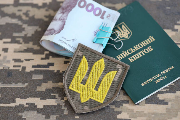 Symbol ukraińskiej armii i legitymacji wojskowej z mnóstwem pieniędzy na mundurze kamuflażu ukraińskiego żołnierza. Koncepcja płatności na rzecz personelu wojskowego lub łapówki za odroczenie służby wojskowej - Zdjęcie, obraz