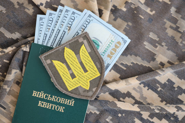 Σύμβολο του ουκρανικού στρατού και στρατιωτική ταυτότητα με πολλά χρήματα στη στολή καμουφλάζ ενός ουκρανού στρατιώτη. Έννοια της πληρωμής στο στρατιωτικό προσωπικό ή δωροδοκία για αναβολή της στρατιωτικής θητείας - Φωτογραφία, εικόνα