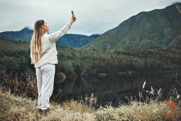 Woman, selfie and forest, mountains or nature in search of network, connection or internet outdoor. Young person camping, travel or hiking with adventure photography or mobile for location by lake. - Photo, Image