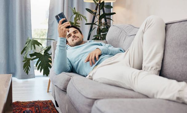 Человек, телефон и отдых на диване для социальных медиа, общения или развлечений в гостиной дома. Мужчина лежит на диване в гостиной с мобильным смартфоном для онлайн-приложения или потокового в доме. - Фото, изображение