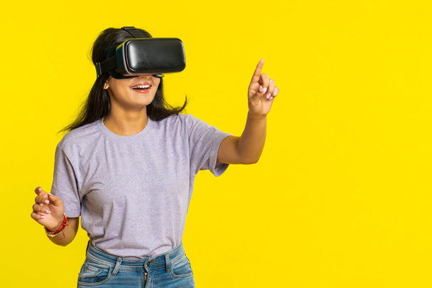 Kulaklık uygulaması kullanarak simülasyon oyunu oynayan heyecanlı Hintli genç kadın. Sanal gerçeklik 3D 360 videosu izliyoruz. VR gözlüklü Arap kız sarı arka planda izole edilmiş. Gelecek teknolojisi - Fotoğraf, Görsel