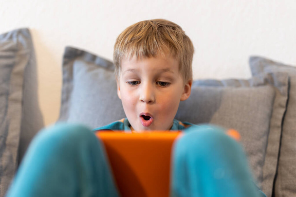 Портрет милый очаровательный блондин маленький мальчик нравится сидеть на домашнем диване серфинг веб-игры или смотреть ТВ-шоу фильмы с помощью планшета. Концепция использования детских умных технологий. Детская гаджетная зависимость. - Фото, изображение