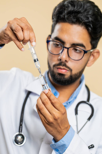 Indiai orvos kardiológus férfi tartja fecskendő tű és ampulla cső orvosi vakcina gyógyszer kezelés injekció használatra kész. Védőoltás. Arab tudós fickó bézs háttérrel. Függőleges - Fotó, kép