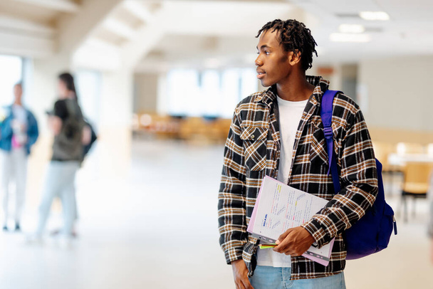Porträt eines jungen schwarzen Mannes, College-Student, der mit Zetteln in der Hand auf dem Flur des Campus steht. Bildung, Vielfalt und Inklusion - Foto, Bild