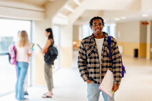 Портрет молодого чорного чоловіка, студента коледжу, що стоїть в коридорі кампусу з нотами в руці. Освіта, різноманітність та включення - Фото, зображення