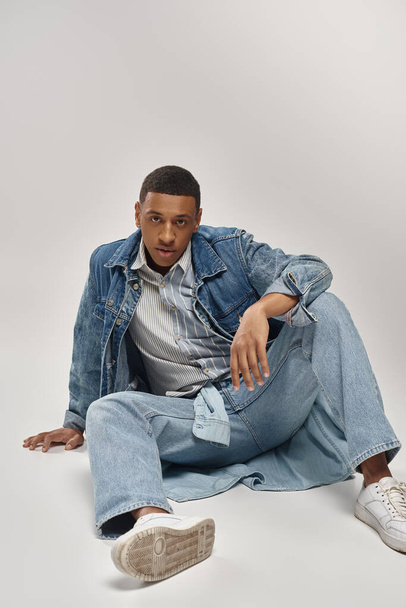 good looking young african american man in trendy denim outfit sitting on floor, fashion concept - Photo, Image
