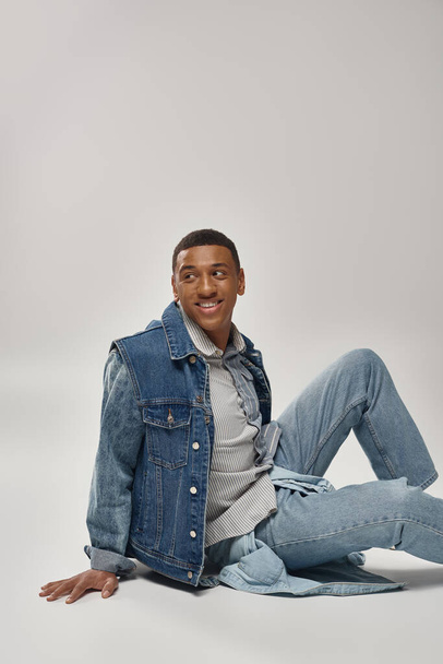 εμφανίσιμος νεαρός αφροαμερικάνος με μοντέρνα denim εμφάνιση κάθεται στο πάτωμα, έννοια της μόδας - Φωτογραφία, εικόνα