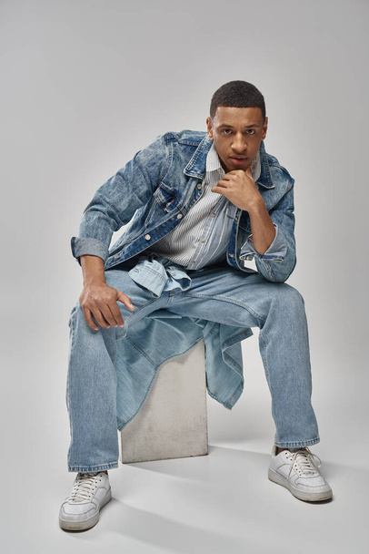 good looking african american male model in denim outfit sitting on white cube, fashion concept - Photo, Image