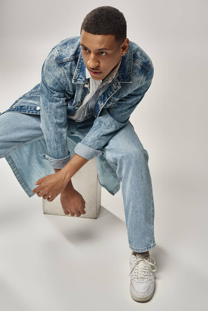 modelo masculino afroamericano atractivo en traje de mezclilla sentado en cubo blanco, concepto de moda - Foto, Imagen