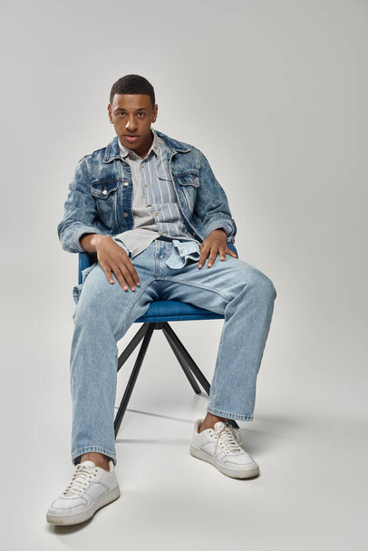 красивый африканский американец в стильном джинсовом наряде сидит на синем стуле, концепция моды - Фото, изображение