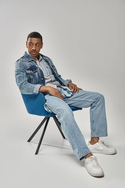 hombre afroamericano guapo en traje de mezclilla elegante sentado en silla azul, concepto de moda - Foto, Imagen