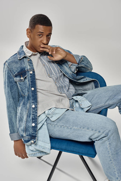 青い椅子に座っているスタイリッシュなデニム服のハンサムなアフリカ系アメリカ人男性,ファッションコンセプト - 写真・画像