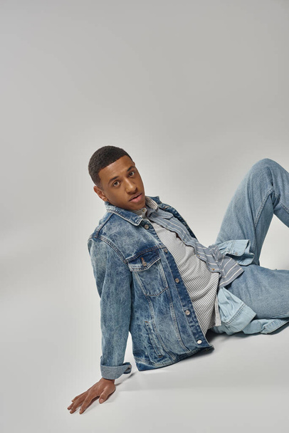 εμφανίσιμος νεαρός αφροαμερικάνος με μοντέρνα denim εμφάνιση κάθεται στο πάτωμα, έννοια της μόδας - Φωτογραφία, εικόνα
