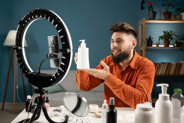 Male beauty influencer sitting in front of smartphone set on tripod, streaming from home, promoting cosmetics. Copy space - Photo, Image