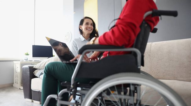 Χαμογελαστή γυναίκα επικοινωνεί με τη γυναίκα που κάθεται σε αναπηρική καρέκλα με φορητό υπολογιστή. Επικοινωνία με άτομα με ειδικές ανάγκες - Φωτογραφία, εικόνα