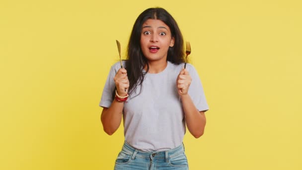 Ready to eat. Hungry Indian woman waiting for serving dinner dishes with with restlessness holding cutlery fork knife will appreciate delicious restaurant meal. Excited girl on yellow background - Footage, Video