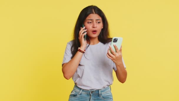 Irritated nervous stressed Indian woman talking screaming on two mobile phones having conversation conflict quarrel complaint dispute discuss solve problem. Arabian girl isolated on yellow background - Footage, Video
