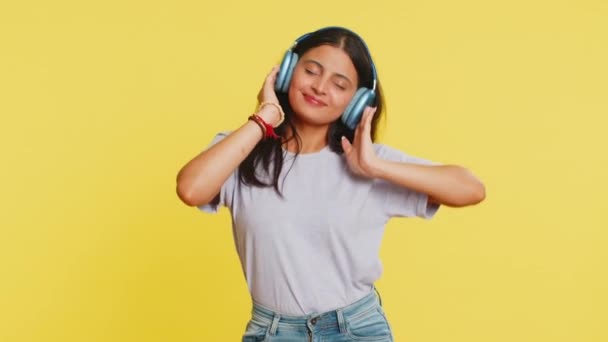 Happy smiling Indian woman listening music in headphones dancing disco fooling around having fun expressive gesticulating hands relaxing chill making funny moves. Hindu girl on yellow background - Footage, Video