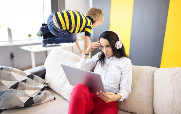 Mujer con auriculares y portátil se sienta en el sofá junto a los mimos de los niños. Cómo combinar el trabajo remoto en el hogar y el concepto de los niños. - Foto, Imagen