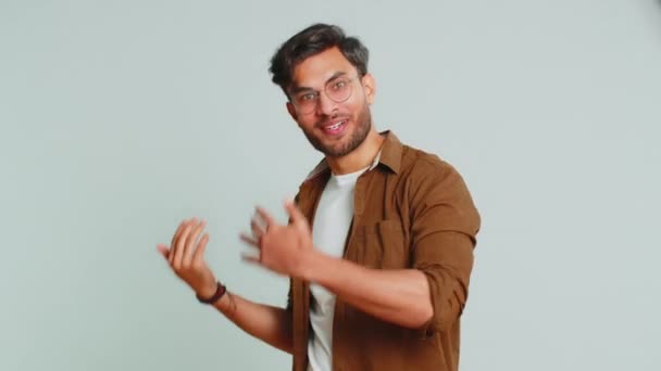 Fiatal indiai férfi barátságos mosollyal a kamerába, integető kézzel üdvözlő hello vagy viszlát üdvözlő meghívó vendégszerető kifejezés. Arab hindu srác elszigetelt szürke stúdió háttér - Felvétel, videó