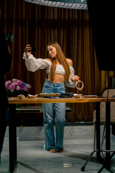 портрет красивої молодої рукодільниці блогер тримає телефон в руках, роблячи фотографію себе з намистом в руках виробництво ремесел - Фото, зображення