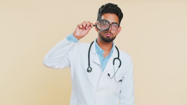 Badacz badacz naukowiec indyjski młody lekarz kardiolog człowiek trzyma szkło powiększające w pobliżu twarzy, wyszukiwanie, analizowanie. Arabian aptekarz facet izolowany na beżowym tle - Materiał filmowy, wideo