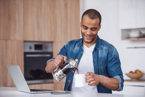 Красивый бизнесмен наливает кофе и улыбается, работая с ноутбуком на кухне - Фото, изображение