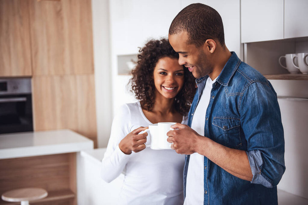 Hermosa pareja está bebiendo café, abrazando y sonriendo mientras descansa en la cocina - Foto, imagen
