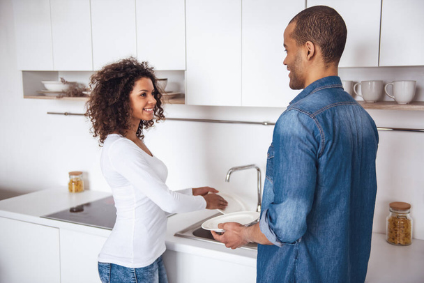 Hermosa pareja está hablando y sonriendo mientras lavan los platos juntos en la cocina - Foto, Imagen