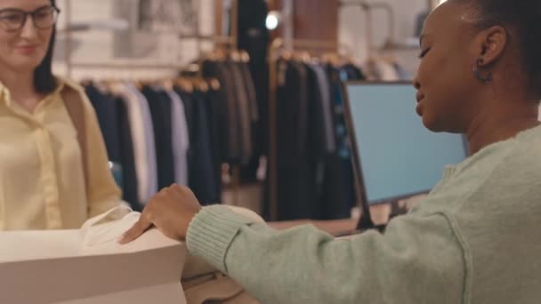 Νεαρή Αφρο-Αμερικανίδα πωλήτρια στέκεται δίπλα στον πάγκο σε κατάστημα πολυτελών σχεδιαστών ρούχων και συσκευάζει ρούχα σε λευκή χάρτινη τσάντα για νεαρή κομψή Καυκάσια γυναίκα στα ψώνια - Πλάνα, βίντεο