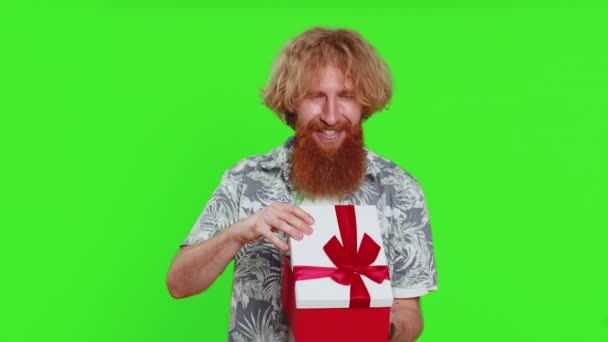 Szczęśliwy biały mężczyzna otwiera pudełko urodzinowe z czerwoną wstążką. Niespodzianka świąteczna. Uśmiechnięty rudy brodaty facet otrzymuje wielki zapakowany prezent świętując izolowane na zielonym tle chroma klucz - Materiał filmowy, wideo