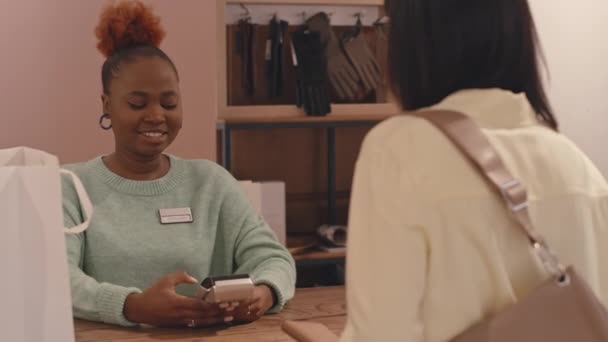 カウンターで若いポジティブな黒人女性ショップアシスタントと小さなおしゃべりをしている女性のお客様,服店で購入しているスマートフォンでお支払い中 - 映像、動画