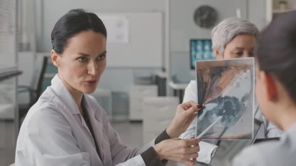Kolmen monietnisen naislääkärin vyötärö, joka tutkii potilaan röntgenkuvaa, kun ryhmäkonferenssi pidetään nykyaikaisessa lääketieteellisessä toimistossa - Materiaali, video