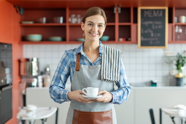 Portrait of smiling caucasian lady barista holding coffee cup and smiling at camera, woman in apron stand at counter in coffee shop - Photo, Image