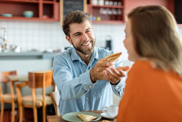 Portrait of smiling european man giving sandwich to his wife, feeding woman while sitting in cafe interior, free space. Nutritious breakfast, snack - Photo, Image
