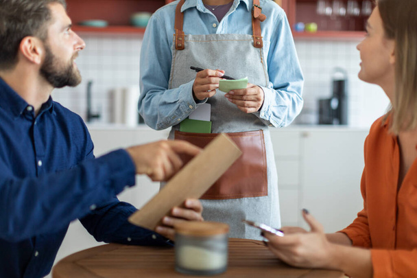 Jeune serveuse portant tablier, prenant ordre dans le café au couple caucasien, homme tenant le menu et parlant au barista - Photo, image