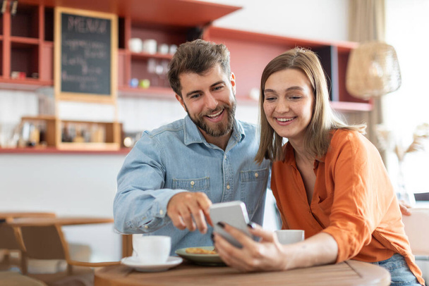 Счастливая европейская пара с помощью смартфона, сидя в кафе вместе в дневное время, серфинг Интернет или покупки в Интернете. Концепция современных отношений - Фото, изображение
