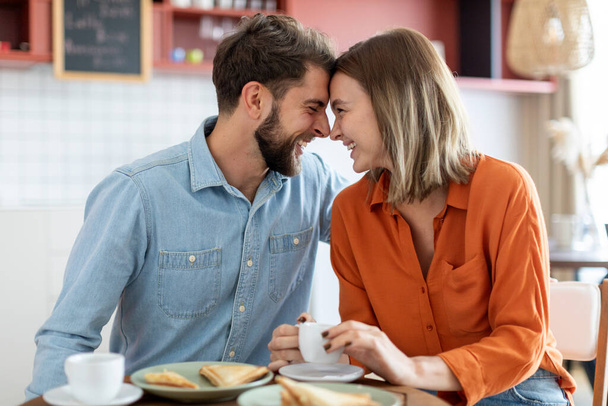 Αγαπώντας το ευρωπαϊκό ζευγάρι απολαμβάνοντας ημερομηνία στην καφετέρια, ο άνδρας και η γυναίκα κάθονται, αγγίζοντας τα μέτωπα και χαμογελώντας, πλευρική άποψη - Φωτογραφία, εικόνα