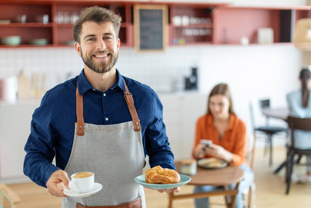 Amistoso camarero masculino sosteniendo taza de café y plato con pastelería, sirviendo orden para el cliente femenino, mirando y sonriendo a la cámara en la cafetería - Foto, Imagen