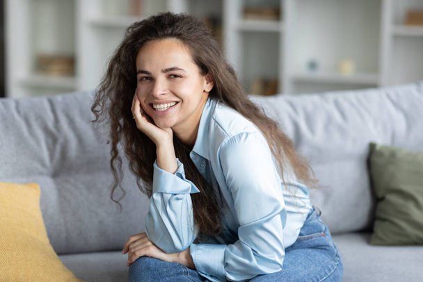 Portrait of beautiful european woman sitting on sofa at home interior, looking and smiling at camera, enjoying domestic leisure, free space - Photo, Image