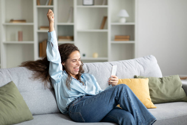 Überglückliche junge Frau hält Smartphone in der Hand und schüttelt vor Freude die Faust, liest gute Nachrichten und Nachrichten, sitzt zu Hause auf der Couch, freier Platz - Foto, Bild