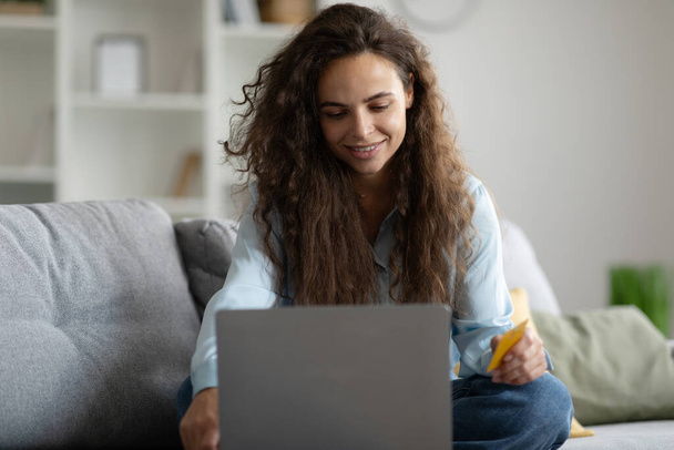 Mujer europea feliz uso de ordenador portátil con tarjeta de crédito, sentado en el sofá en el interior de la sala de estar, compras en línea. Orden y reembolso - Foto, Imagen