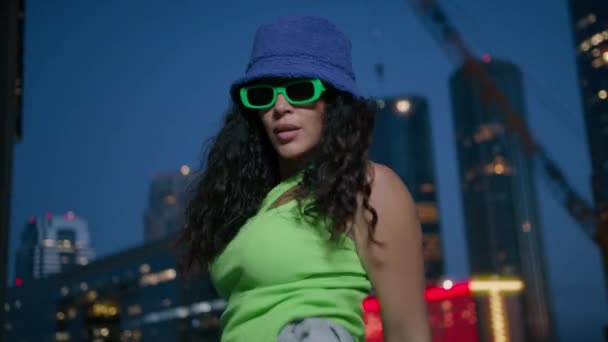 Cool sexy mixed race female modern dancer moves rhythmically 4K. Active african american black woman in neon green top and purple bucket hat dancing by urban lights background in downtown at night - Footage, Video