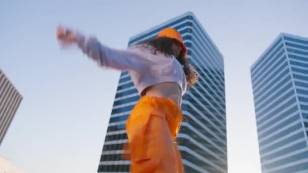 A hip-hop királynő twerket csinál. Breakdancer nő élvezi naplemente 4K. Vidám, többfajú lány sportszerelésben divatos narancssárga nadrágot és vödör sapkát visel, zenére táncol a belvárosban. - Felvétel, videó