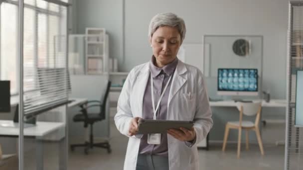 Средний портрет зрелой азиатки-врача в белом халате, позирующей перед камерой с цифровым планшетом в руках в медицинской клинике - Кадры, видео
